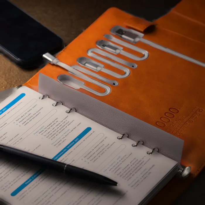 Illustration: the open orange DAYBOOK 360 on a dark table in warm lamplight, a phone beside it plugged into the planner’s built-in USB-C cable, a pen on the page